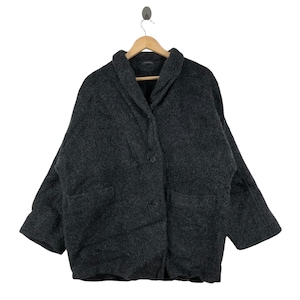 Puede incluir: Un abrigo gris oscuro de gran tamaño con un cuello ancho y dos bolsillos grandes. El abrigo está hecho de una tela texturizada y tiene dos botones. Cuelga de una percha de madera.