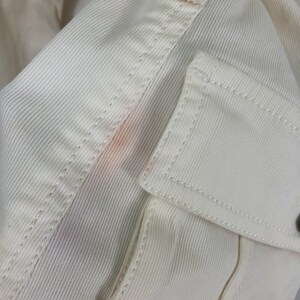 May include: Close-up of a white cotton fabric with a faint pink stain. The fabric has a textured, ribbed pattern and is stitched with a white thread.