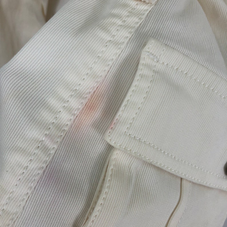May include: Close-up of a white cotton fabric with a faint pink stain. The fabric has a textured, ribbed pattern and is stitched with a white thread.