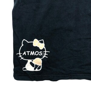 May include: Black t-shirt featuring a white outline of a cat with a yellow bow and a beige ice cream cone. The word "ATMOS" is written in white inside the cat's outline. The t-shirt is made of a soft, dark fabric.