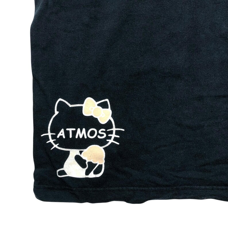 May include: Black t-shirt featuring a white outline of a cat with a yellow bow and a beige ice cream cone. The word "ATMOS" is written in white inside the cat's outline. The t-shirt is made of a soft, dark fabric.