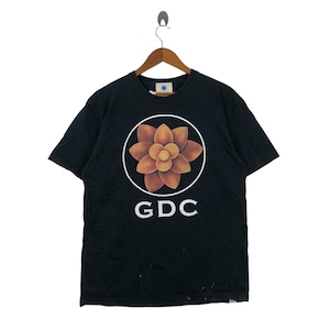 Puede incluir: Camiseta negra con un diseño circular de una flor estilizada en tonos naranja y amarillo, rodeada por un anillo blanco. Las letras "GDC" están impresas en blanco debajo. La camiseta está colgada de una percha de madera.