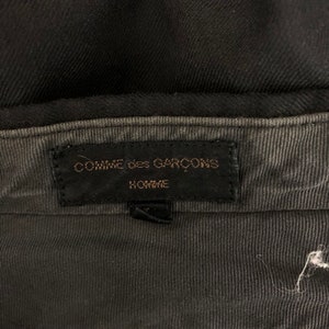 May include: Close-up of a black garment's interior, showcasing a label with the text "COMME des GAR&Ccedil;ONS HOMME" in gold lettering on a black background. The fabric appears to be a dark gray corduroy.