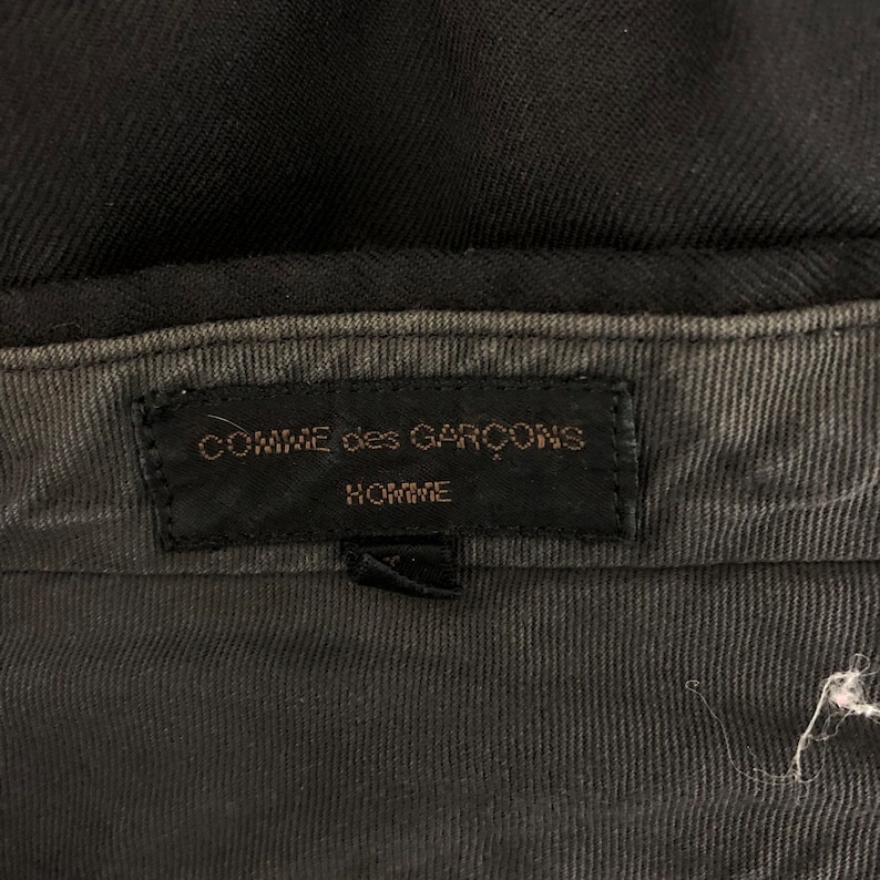 May include: Close-up of a black garment's interior, showcasing a label with the text "COMME des GAR&Ccedil;ONS HOMME" in gold lettering on a black background. The fabric appears to be a dark gray corduroy.