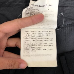 May include: A close-up of a tag from a garment, featuring text in English and Japanese. The tag indicates the item is made in Japan from 100% wool and provides care instructions, including dry clean only. The brand name is visible.