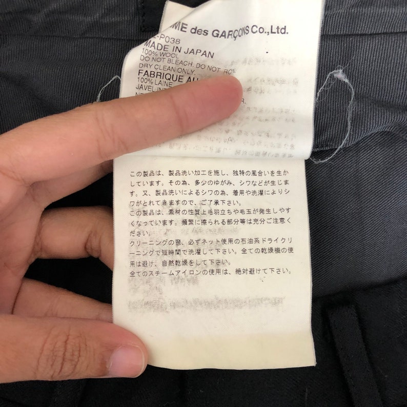 May include: A close-up of a tag from a garment, featuring text in English and Japanese. The tag indicates the item is made in Japan from 100% wool and provides care instructions, including dry clean only. The brand name is visible.