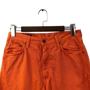 Op de afbeelding: Een paar oranje spijkerbroeken met een bruine houten hanger. De spijkerbroek heeft een klassieke vijf-zak stijl en een knoopsluiting.