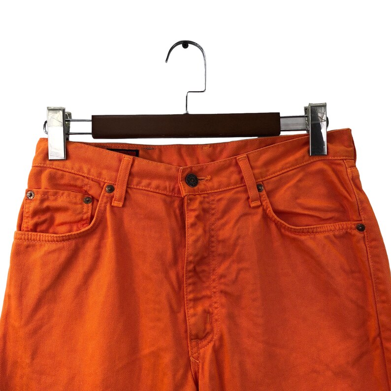 Op de afbeelding: Een paar oranje spijkerbroeken met een bruine houten hanger. De spijkerbroek heeft een klassieke vijf-zak stijl en een knoopsluiting.