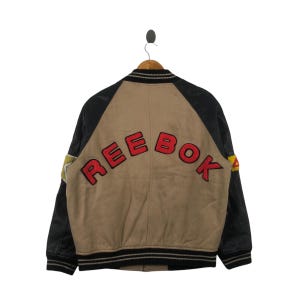 Könnte beinhalten: Eine schwarze und braune Varsity-Jacke mit einem großen roten "Reebok"-Logo auf der Rückseite. Die Ärmel sind aus schwarzem Leder und der Körper ist braun.