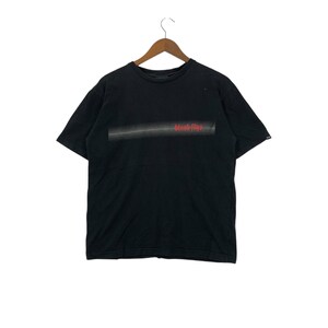 Puede incluir: Camiseta negra con un diseño horizontal y el texto "black flys" en rojo. La camiseta está colgada en una percha de madera sobre un fondo blanco. La camiseta tiene mangas cortas y cuello redondo.