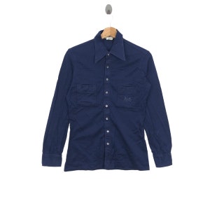 Puede incluir: Una camisa azul marino de manga larga con botones, dos bolsillos en el pecho y un pequeño logotipo bordado en el bolsillo derecho.