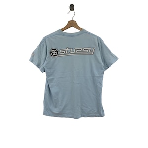May include: A light blue t-shirt with a black and white Stussy logo on the back. The logo is in a stylised font with a black outline and white fill.