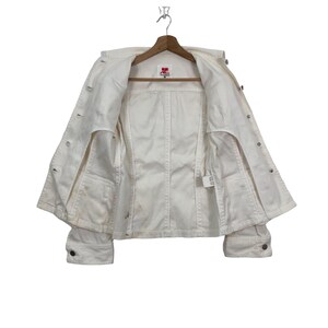 May include: White denim jacket with a button closure and long sleeves. The jacket is unbuttoned and hanging on a hanger. The label on the inside of the jacket reads "JC".