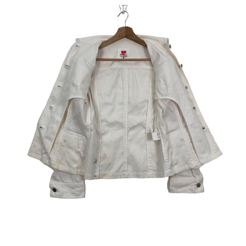 May include: White denim jacket with a button closure and long sleeves. The jacket is unbuttoned and hanging on a hanger. The label on the inside of the jacket reads "JC".