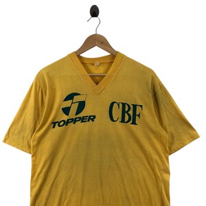 May include: A vintage yellow V-neck t-shirt with the text "TOPPER" and "CBF" in dark green. The shirt has short sleeves and is hanging on a wooden hanger.