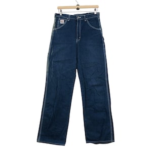 Könnte beinhalten: Dunkelblaue Jeans-Arbeitshose mit weißen Nähten und einem kleinen Aufnäher auf der Vordertasche. Die Hose hängt an einem Holzbügel. Die Jeans hat ein weites Bein und einen klassischen Arbeitskleidungsstil.