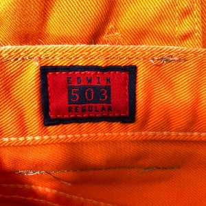 Op de afbeelding: Een close-up van een rood en zwart label met de tekst "EDWIN 503 REGULAR" op een oranje spijkerbroek.