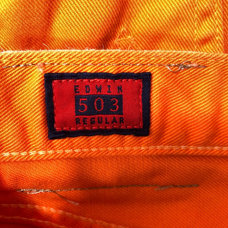 Op de afbeelding: Een close-up van een rood en zwart label met de tekst "EDWIN 503 REGULAR" op een oranje spijkerbroek.
