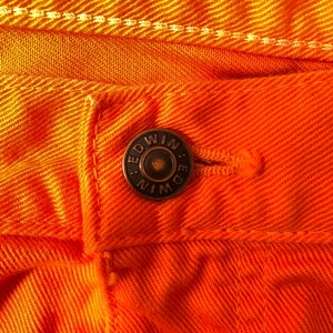 Op de afbeelding: Close-up van een paar oranje spijkerbroeken met een koperen knoop waarop "EDWIN" in hoofdletters staat.