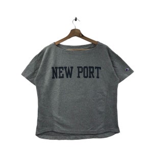 以下が含まれることがあります： グレーの半袖Tシャツ。ネイビーの「NEW PORT」のグラフィックと、右袖に小さなチャンピオンのロゴが入っています。