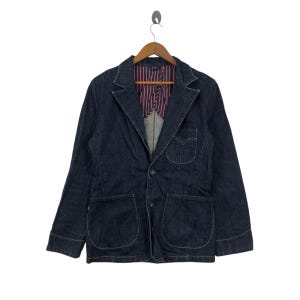 May include: A dark blue denim blazer with a two-button closure, notched lapels, and two front pockets. The interior lining features a striped pattern in red and white. The blazer is hanging on a wooden hanger.