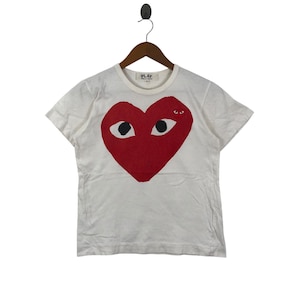 Può includere: T-shirt bianca con un grande cuore rosso con occhi neri. La maglietta ha maniche corte e girocollo. La parola "PLAY" è stampata sul collo. La maglietta è appesa a una gruccia di legno.