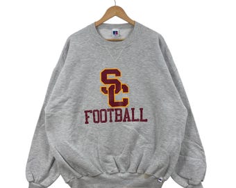 Puede incluir: Sudadera gris jaspeada con el texto "SC FOOTBALL" en granate y dorado. La sudadera está colgada de una percha de madera. Tiene cuello redondo y mangas largas.