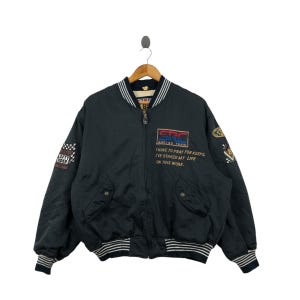 Może przedstawiać: Czarna kurtka bomberowa z zamkiem błyskawicznym i prążkowanymi ściągaczami. Kurtka ma kilka naszywek haftowanych, w tym jedną z napisem "SRC Racing Team" i drugą z napisem "I have to pray for keeps, I've staked my life on this work."