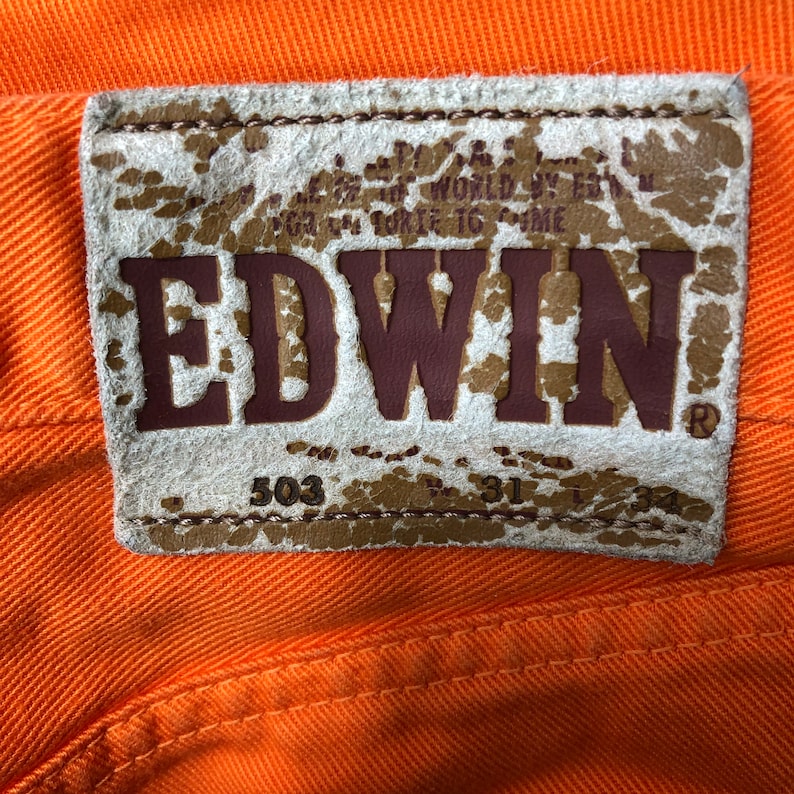 Op de afbeelding: Een close-up van een bruin leren patch met het woord "EDWIN" in bruine letters. De patch heeft ook de nummers "503" en "31" erop gedrukt.