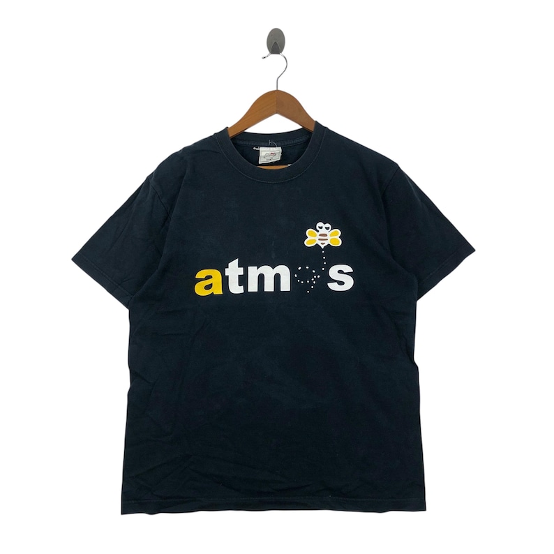 May include: Black t-shirt with the word "atmos" in yellow and white, and a cartoon bee graphic. The t-shirt is hanging on a wooden hanger.