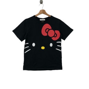 K&ouml;nnte beinhalten: Schwarzes T-Shirt mit einem wei&szlig;en Hello Kitty-Motiv und einer roten Schleife. Das Shirt hat einen Rundhalsausschnitt und kurze &Auml;rmel.