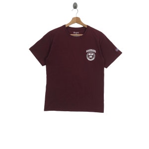 Puede incluir: Camiseta de manga corta color burdeos con un logotipo blanco de Champion en la manga izquierda y un logotipo blanco de la Universidad de Harvard en la parte delantera.
