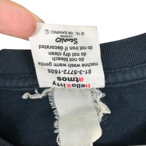 May include: Close-up of a black t-shirt tag with the Hello Kitty and Atmos logos in red. The tag includes care instructions and the Sanrio license information.