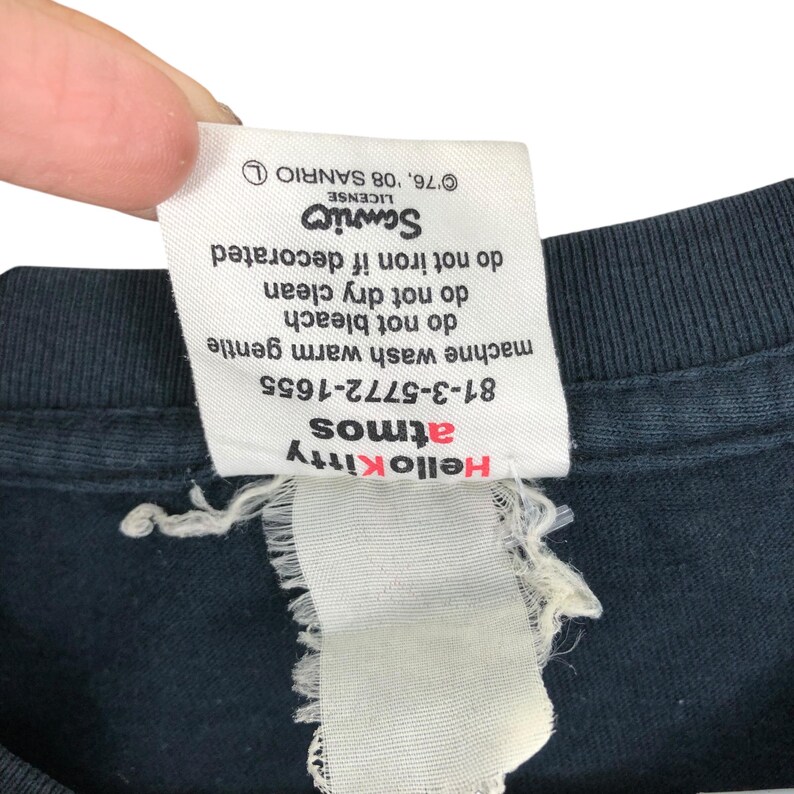 May include: Close-up of a black t-shirt tag with the Hello Kitty and Atmos logos in red. The tag includes care instructions and the Sanrio license information.