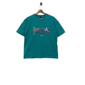 以下が含まれることがあります： 緑がかった青色のTシャツに、一緒に立っている人々のグループのグラフィックプリントが施されています。グラフィックの下には「SECTION 8」というテキストが印刷されています。