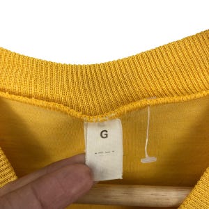 May include: Close-up of a yellow shirt with a ribbed neckline. A white tag with the letter "G" is attached to the shirt. The shirt is hanging on a wooden hanger.