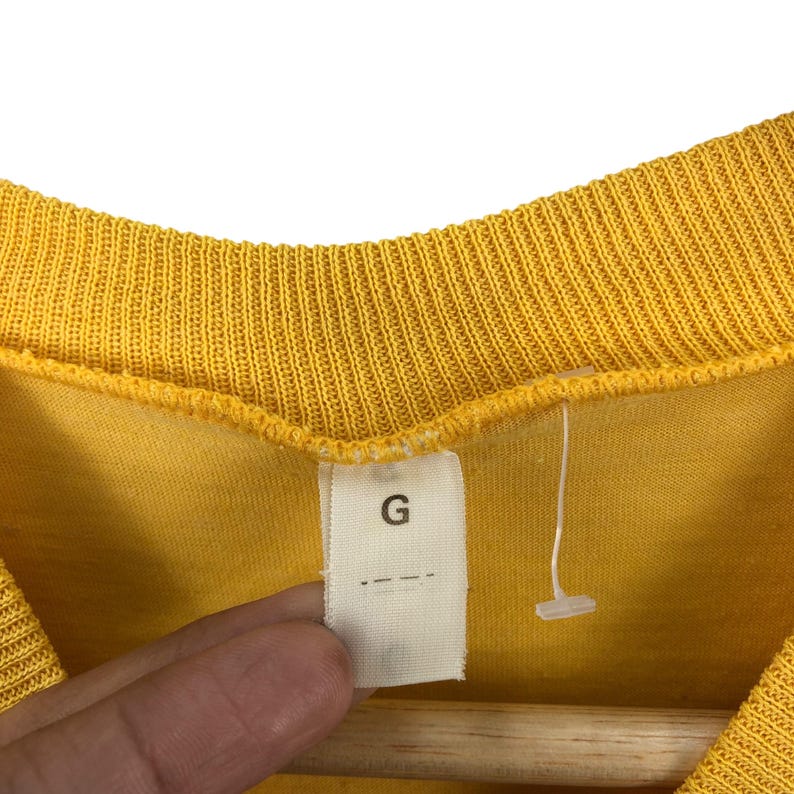 May include: Close-up of a yellow shirt with a ribbed neckline. A white tag with the letter "G" is attached to the shirt. The shirt is hanging on a wooden hanger.