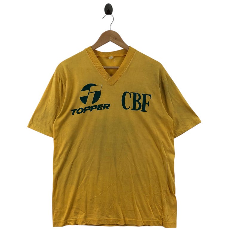 May include: Mustard yellow V-neck t-shirt with the text "TOPPER" and "CBF" in dark green. The shirt has short sleeves and is hanging on a wooden hanger.