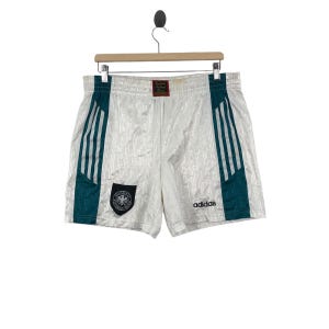 Pode incluir: Calções desportivos brancos com riscas e detalhes em verde-azulado e branco. Os calções apresentam o logótipo Adidas e um emblema nacional alemão. Os calções estão pendurados num cabide de madeira.