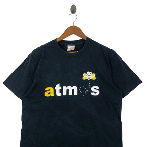 May include: Black t-shirt with the word "atmos" in yellow and white, and a cartoon bee graphic. The t-shirt is hanging on a wooden hanger.