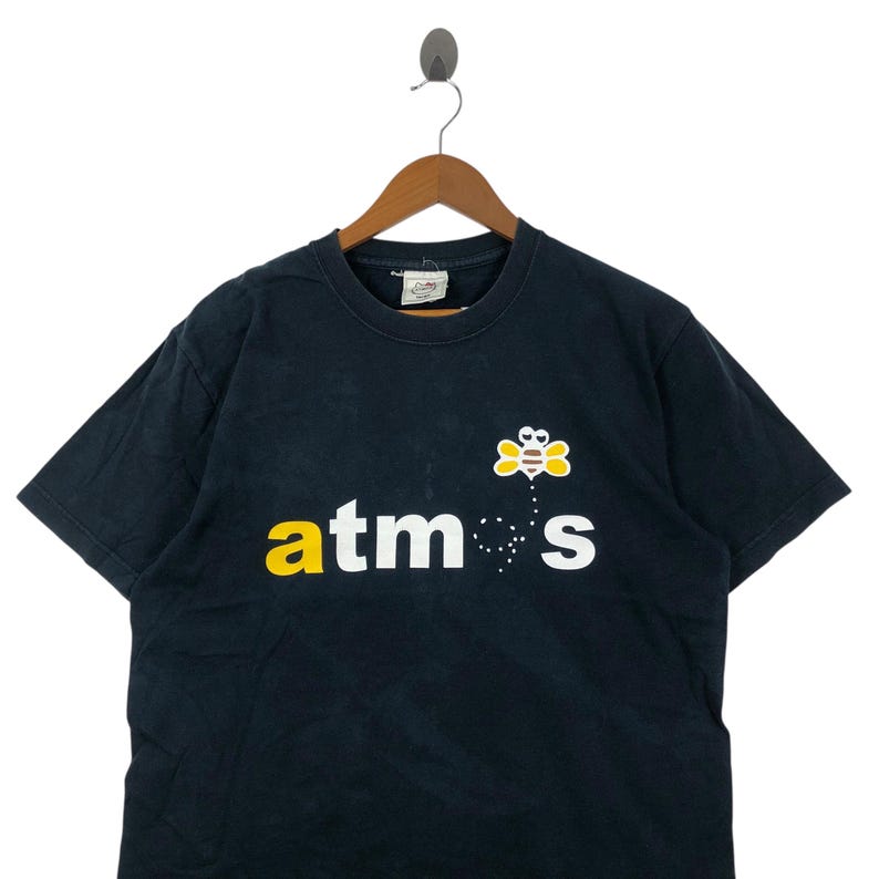 May include: Black t-shirt with the word "atmos" in yellow and white, and a cartoon bee graphic. The t-shirt is hanging on a wooden hanger.