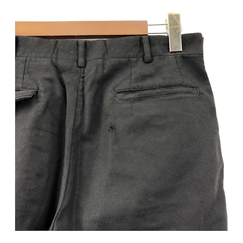May include: Close-up of black pants hanging on a wooden hanger. The pants have a buttoned back pocket and a small hole near the pocket. The pants are made of a solid black fabric.