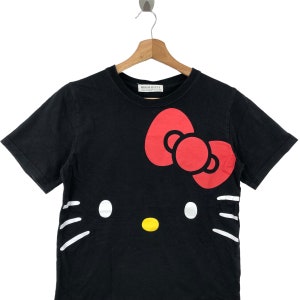 K&ouml;nnte beinhalten: Schwarzes T-Shirt mit kurzen &Auml;rmeln und einem wei&szlig;en Hello-Kitty-Motiv. Das Motiv zeigt Hello Kittys charakteristisches rotes Schleifenband und wei&szlig;e Schnurrhaare.