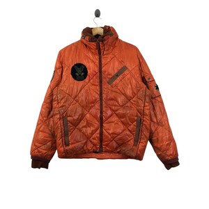 May include: Orange quilted jacket with brown accents, a high collar, and a front zipper. Features a patch on the chest with the text "USOS" and a circular emblem. The jacket has a diamond pattern and brown ribbed cuffs.