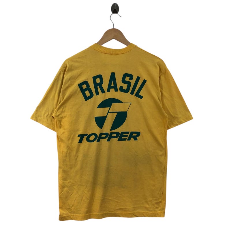 May include: A yellow t-shirt featuring the word "BRASIL" in green, arched above a green and white logo, with "TOPPER" below. The shirt is suspended from a wooden hanger.