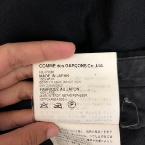 May include: A close-up of a white tag with black text from Comme des Gar&ccedil;ons Co., Ltd. The tag reads "MADE IN JAPAN" and "100% WOOL." Care instructions are also printed on the tag.