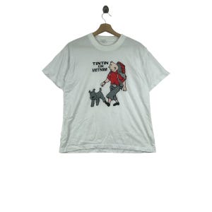 May include: White t-shirt featuring a cartoon illustration of a character with a backpack, a dog, and the text "TINTIN IN VIETNAM". The graphic is in red, black, gray, and tan. The shirt has short sleeves and a crew neck.