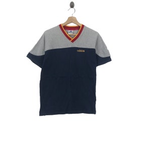 May include: A navy blue and grey short-sleeved t-shirt with a red and yellow v-neck collar. The shirt has the word "adidas" embroidered on the front in yellow.