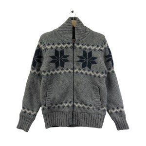 Könnte beinhalten: Ein grauer Strickpullover mit Reißverschluss und einem Schneeflockenmuster in Marineblau. Der Pullover hat zwei Taschen und einen gerippten Saum und Bündchen.