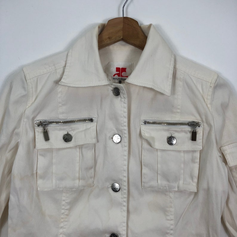 May include: A white cotton jacket with a red logo that says "Couture" and silver zipper accents. The jacket has a button closure and two flap pockets with zipper closures.